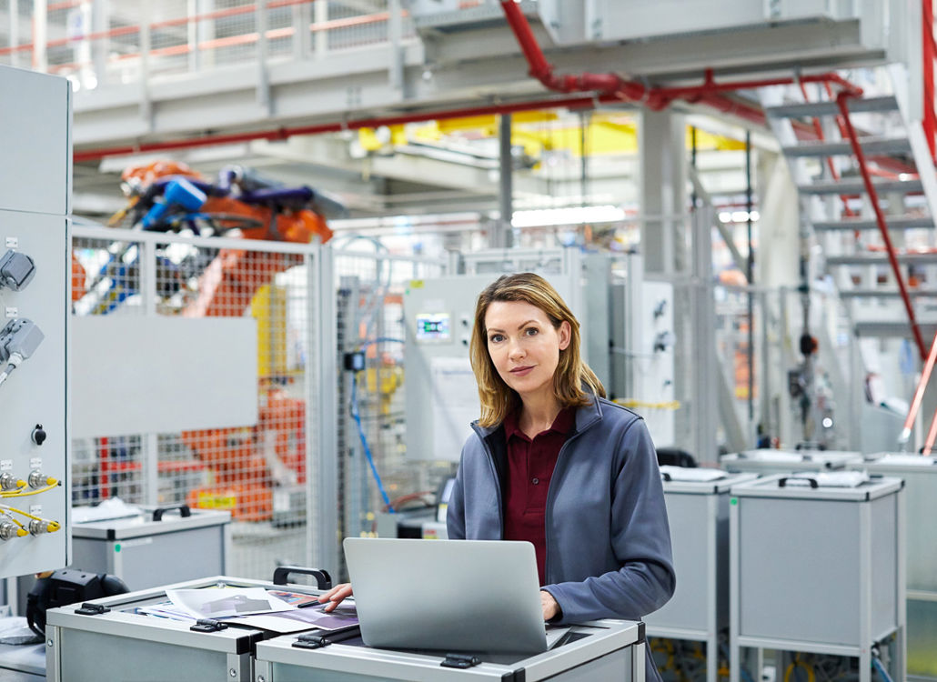 A confident female engineer working on a laptop in a modern industrial facility, monitoring automated production systems.